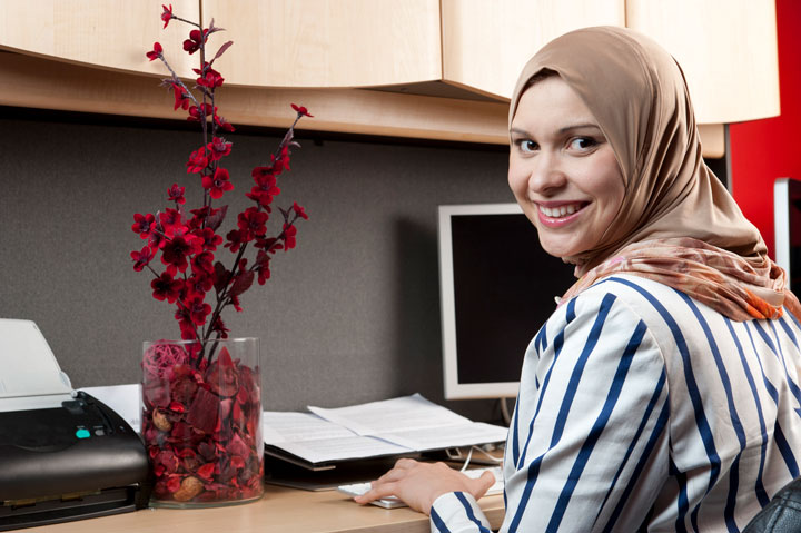 A women sits at her desk turning her head behind to smile at the camera. She wears a hijab and a stripped long sleeve shirt.