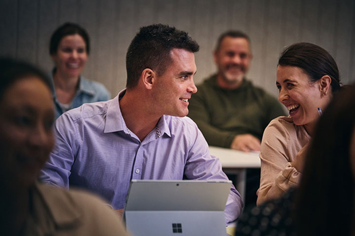 A student with his laptop open engages with a laughing fellow student in a face-to-face class.