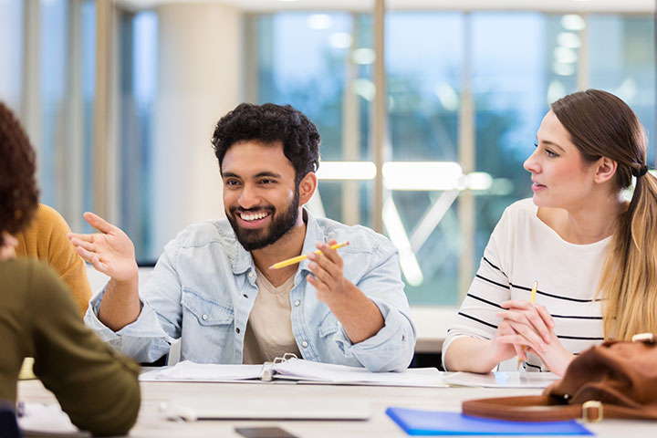 People sit at a desk with their study materials in front of them and pencils in their hands. One man gestures openly with his hands and a broad smile on his face while the woman n his left turns to face him, hands clasped.