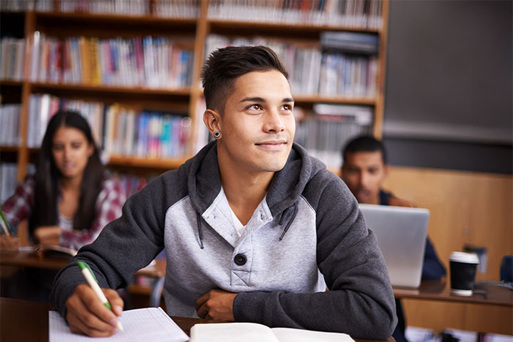A young man sits in a classroom, writing in a notepad while smiling up at his teacher. There are two other students and a bookshelf in the background.