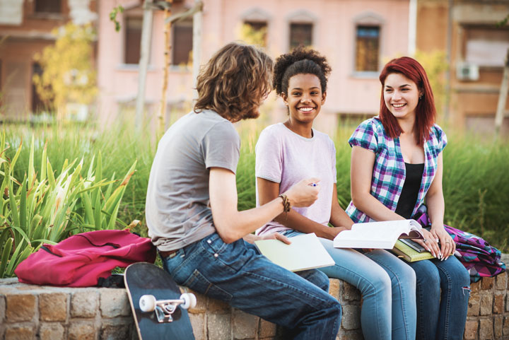 Three students sit on a brick wall on a TAFE NSW campus outdoors. They talk to each other while holding books, pens and notepads. They have backpacks and skateboards with them.