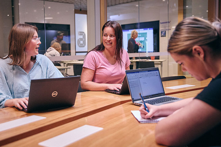 Three female students work opposite each other on laptops and note pads at a shared, contemporary and well-lit desk space. Other students are seen in a separate study area through a glass office wall behind them.