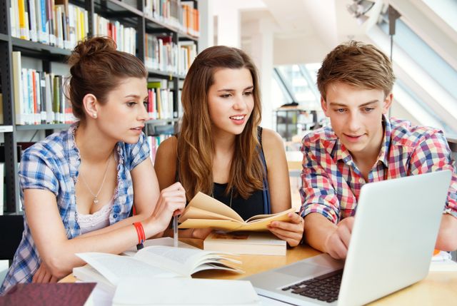 Three students work together in a library, looking at a laptop and reading books. Stacks of books on shelves are visible in the background.