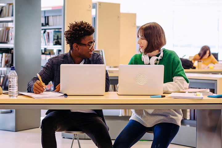 Two students discussing while using their laptops in a library