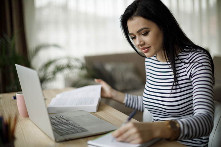 A young student wearing a striped top writes in her notebook and reviews a book, with her laptop out.