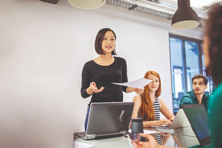 People gather around a table. One woman is standing and gesturing as she talks to the room, while holding a piece of paper.