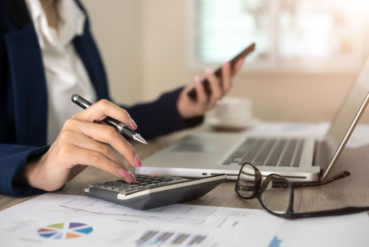 A close-up image of a woman at a desk with her laptop. She’s holding a phone in one hand and a pen in the other. There’s a calculator, a pair of glasses and some worksheets on the desk beside her.