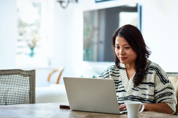 A women in a striped knit jumper sits are her dinner table with her laptop open and a mug beside it.