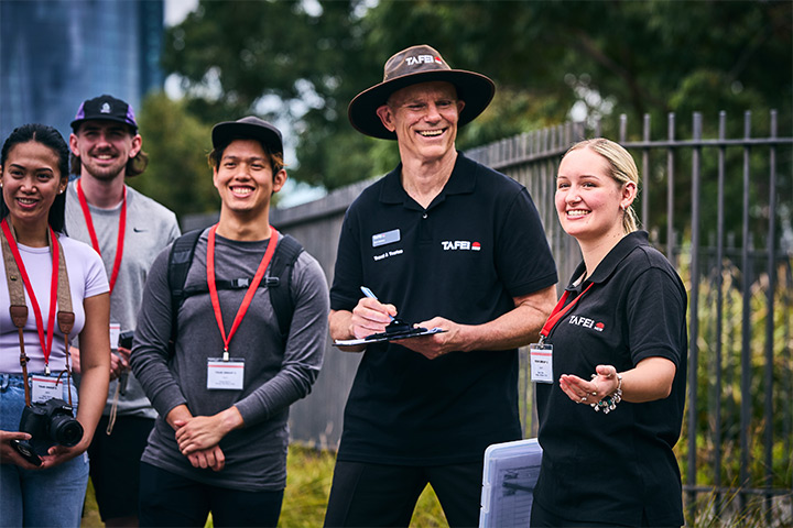 Tourism and travel management teacher leads a group of students on an outdoor field tour, standing near an iron fence while referencing notes on a clipboard as students listen and observe.