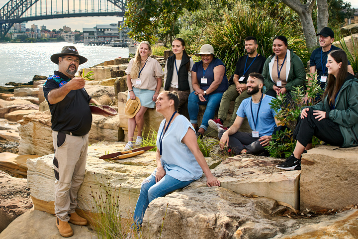 Aboriginal TAFE NSW teacher wearing a black hat and holding a plant, leading a tour group seated on rocks along the banks of Darling Harbour, with the Sydney Harbour Bridge visible in the background.