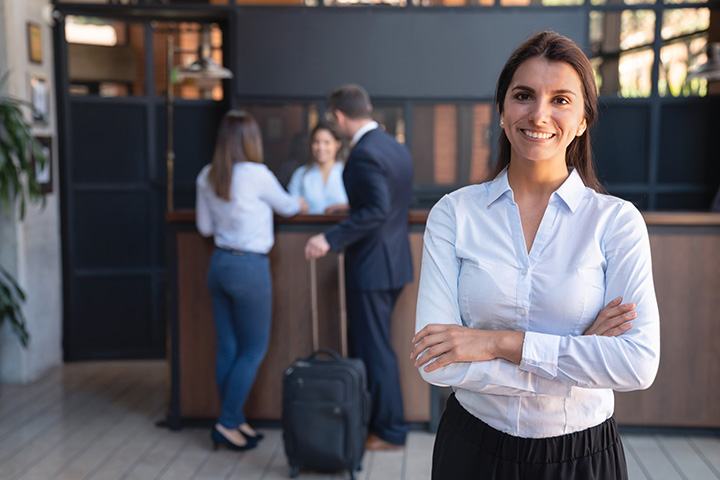 A hotel manager smiles for a photo while a guest checks in at the reception desk in the background.