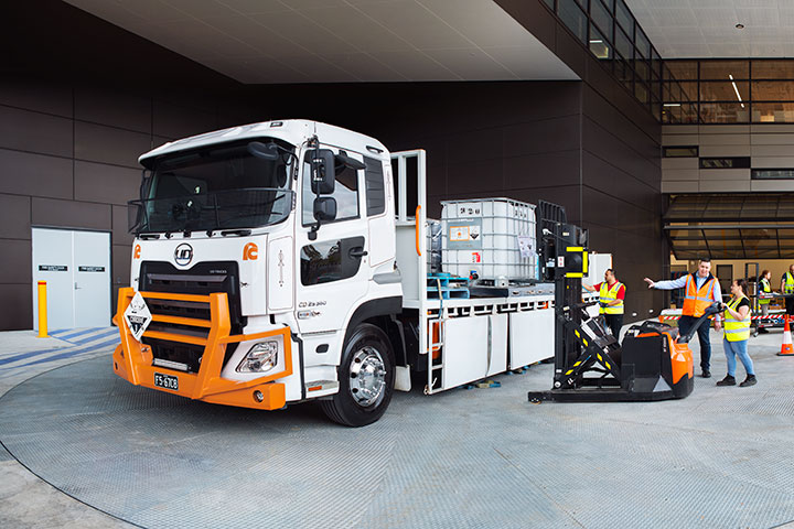 TAFE NSW logistics training scenario featuring a white and orange truck with containers on a flatbed, individuals in high-visibility vests and hard hats operating a forklift and engaging in transportation and logistics activities at an industrial training facility.