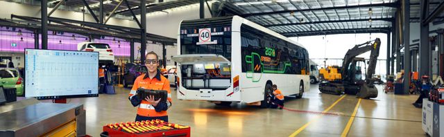Female student in high-visibility orange and navy blue workwear. She wears PPE including safety glasses and stands confidently in front of a modern electric vehicle bus, in an industrial training environment.
