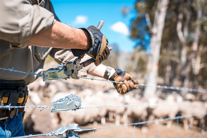 Close-up of farmer's hands working on a steel fence with blurred sheep in the background, suggesting a rural farm setting.