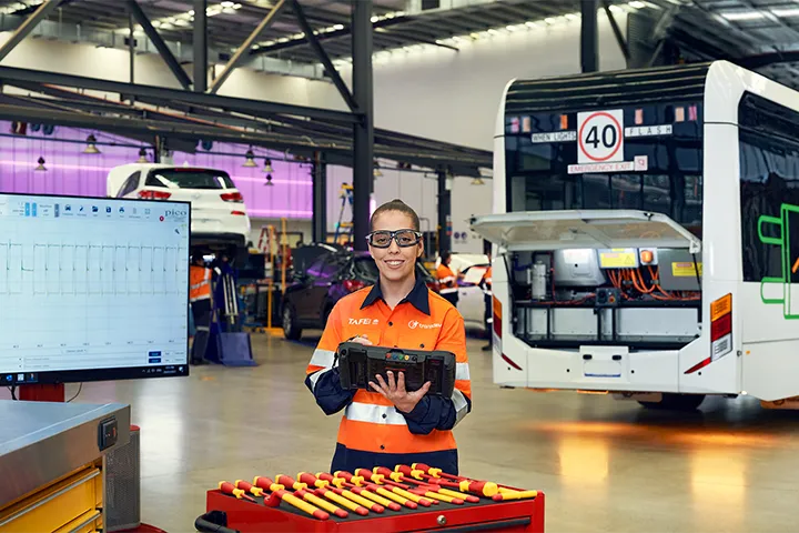 EV bus mechanic student stands in a large automotive workshop wearing high‑visibility protective clothing, holding diagnostic equipment beside a workstation with insulated tools. Behind the student, an electric bus with an open rear compartment is being serviced, while other heavy vehicles and machinery are positioned throughout the workshop.