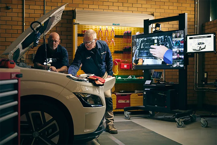 Automotive EV student and teacher work together on an electric vehicle in a workshop, examining components under the open bonnet while referencing instructions on two large display screens. A wall of organised tools and diagnostic equipment is visible in the background.