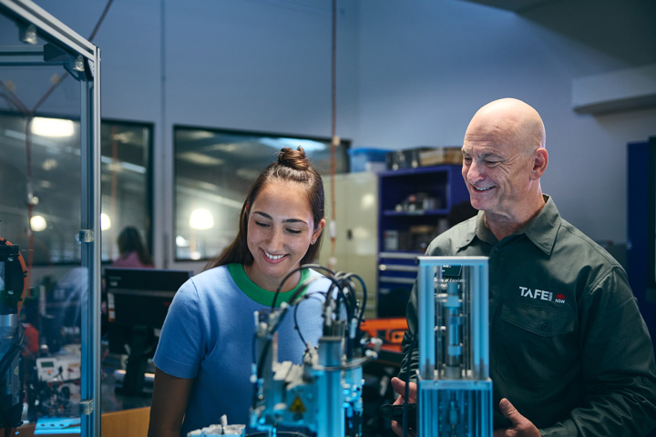 TAFE NSW advanced manufacturing teacher and student smile while examining automated manufacturing equipment in a training workshop, with precision machinery, safety enclosures, and industrial tools visible as part of Centres of Excellence advanced manufacturing training.