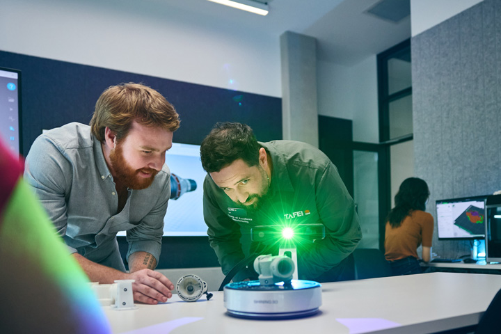 TAFE NSW students examine a component using a handheld 3D scanning device emitting a green light onto a mechanical part on a workstation, with a digital display showing a 3D model in the background inside a modern manufacturing training lab.