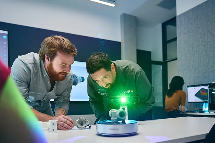 TAFE NSW students examine a component using a handheld 3D scanning device emitting a green light onto a mechanical part on a workstation, with a digital display showing a 3D model in the background inside a modern manufacturing training lab.