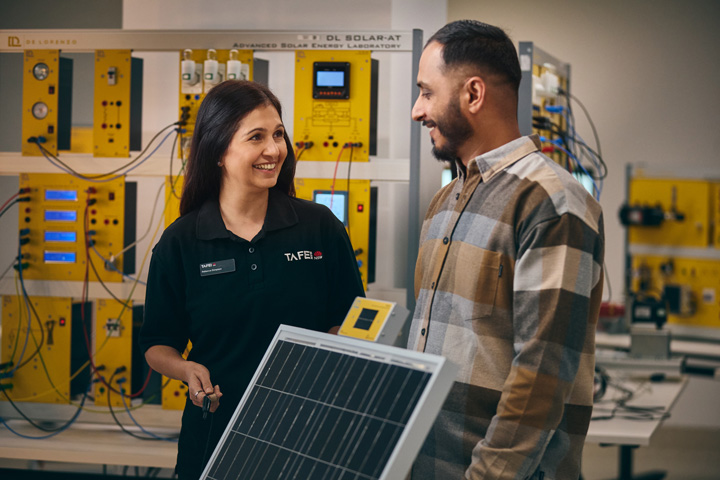 TAFE NSW teacher and student stand in a renewable energy training lab holding a solar panel and control unit, smiling while discussing equipment, with yellow solar training systems and wiring visible behind them as part of Centres of Excellence renewable manufacturing training.