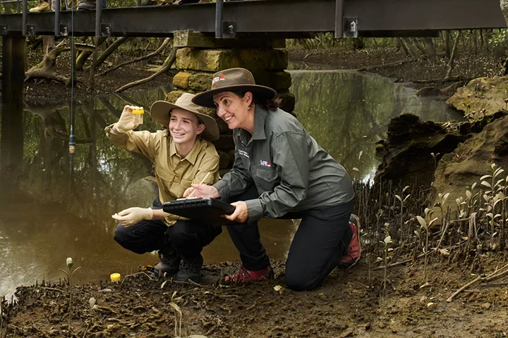 A teacher and student bend down at the edge of a creek, carefully collecting water samples in containers for a science experiment.
