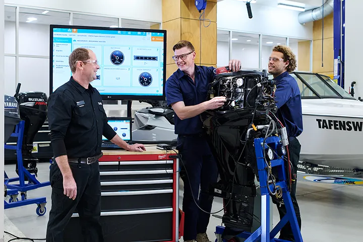 A male marine mechanic in a classroom workshop setting, guiding two students as they work together to troubleshoot and repair an outboard. A screen with instructional materials is visible in the background.