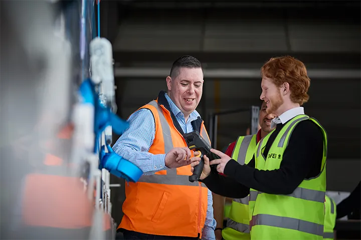 Three people in high-visibility vests on a factory floor. A logistics teacher, wearing an orange vest, points out features of equipment to two students wearing yellow vests.