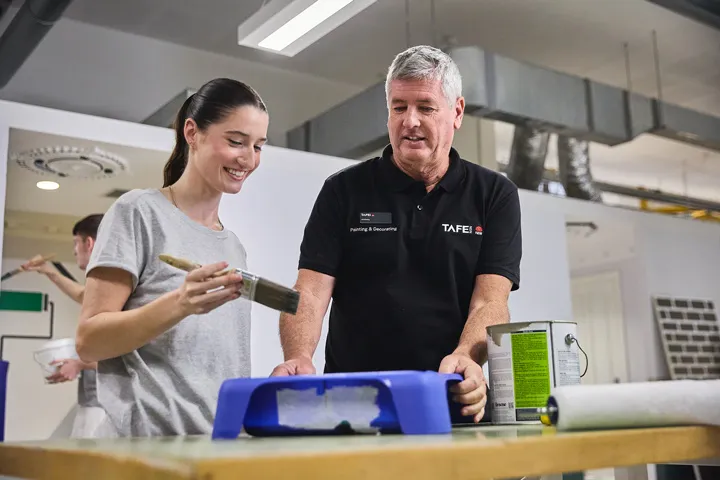 Painting and decorating student applies paint with a brush while a TAFE NSW teacher provides guidance at a workbench, with paint trays, rollers, and tins visible in a well‑lit training workshop during hands‑on practical instruction.