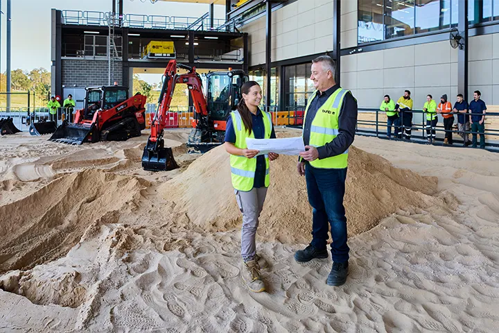 A female student works with the teacher in a civil construction workshop with a bulldozer and excavator in the background.