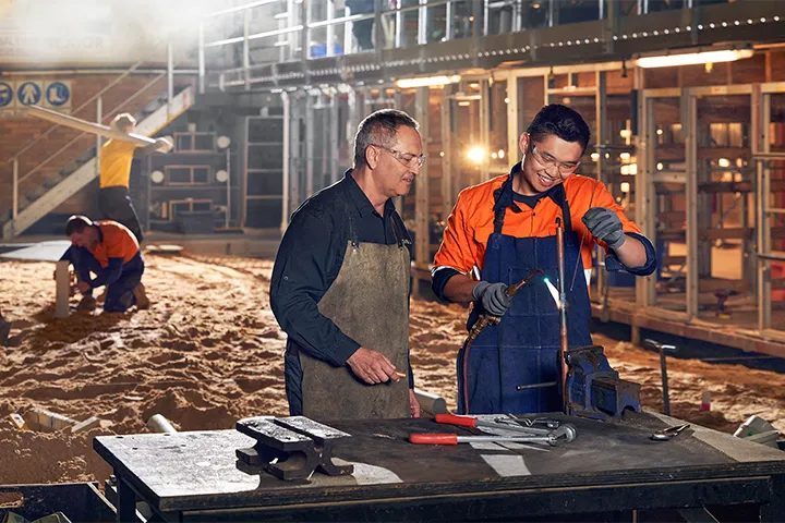 Teacher and student working together on a construction site, skillfully welding a pipe to enhance practical knowledge in the field of construction and welding.