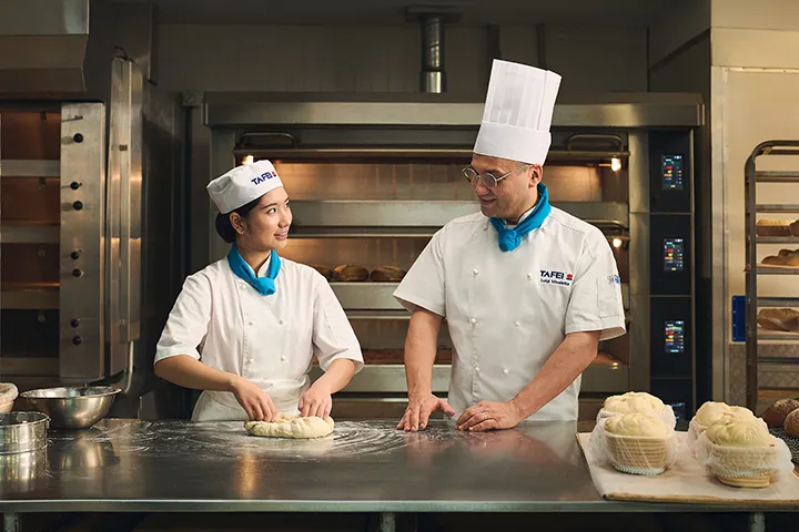A baking student and their instructor share a smile while kneading dough during a bread-making lesson.
