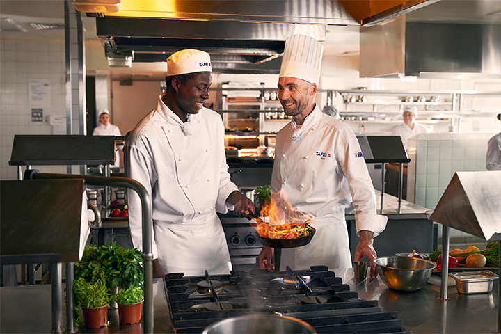 Teacher and student chef collaborating in a bustling commercial kitchen, honing skills and creating culinary masterpieces together.