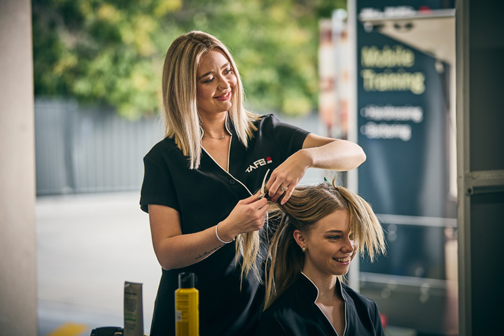 Hairdressing student practices sectioning and combing hair on a client during a training session, wearing a TAFE NSW uniform with hair products and tools arranged on the workstation in an outdoor mobile training setup.