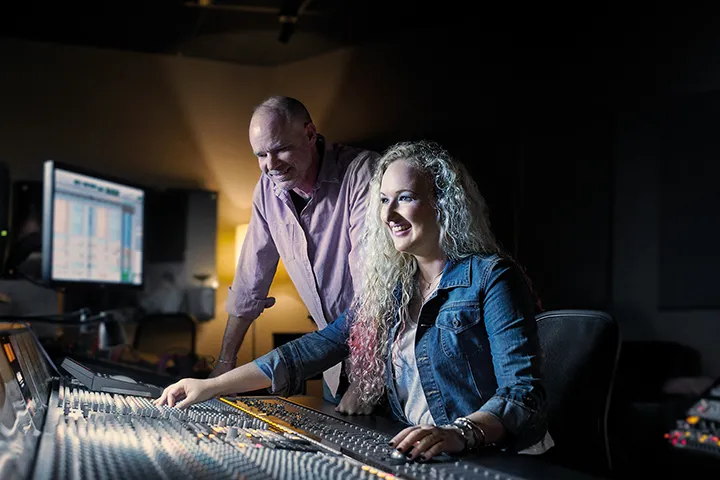 A music engineering teacher and student collaborating on a soundboard in a professional studio environment.