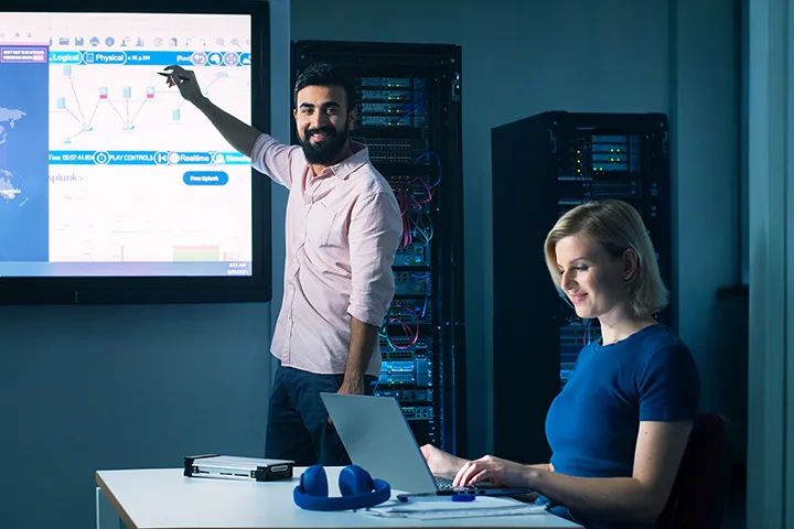 A cyber security teacher pointing at a digital board while a student works on a laptop in a classroom setting.