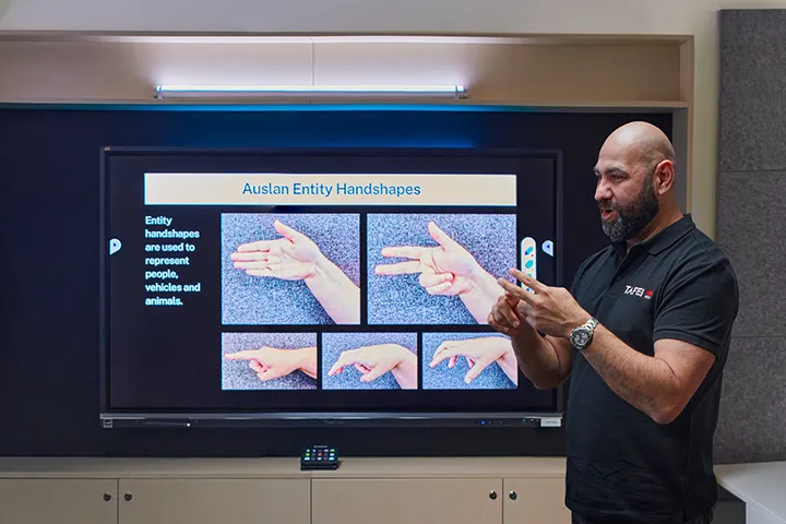 Male teacher teaching Auslan, gesturing while a screen behind him shows a range of different Auslan handshapes and signs.