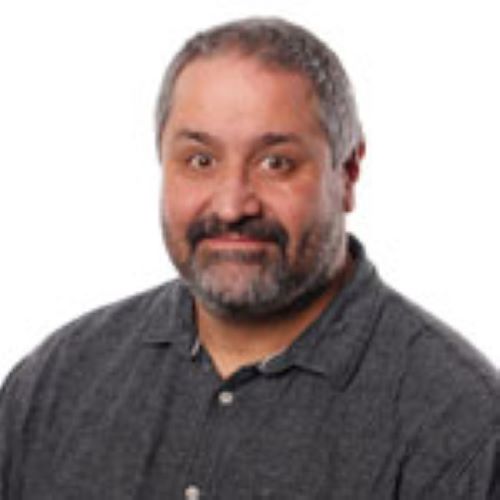 A headshot image of Andy Jenkinson smiling at the camera. He is wearing a dark grey collared shirt and has a salt-and-pepper moustache and beard. 