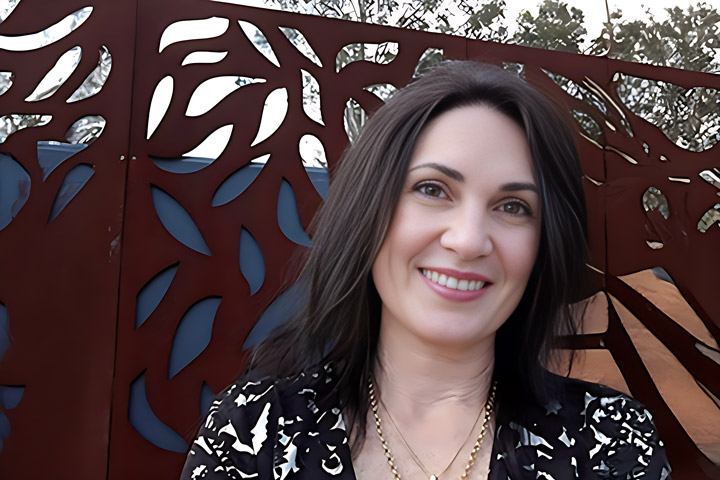 Dee Wardle, Early Childhood teacher at TAFE NSW, stands in a garden beside a decorative metal gate. She is dressed in a black and white patterned dress and gold jewellery.