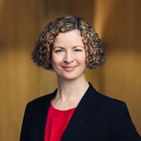 A headshot of Jessica Tilley, smiling and looking directly at the camera. She has short, curly bronde hair, and is wearing a black blazer over a red top.
