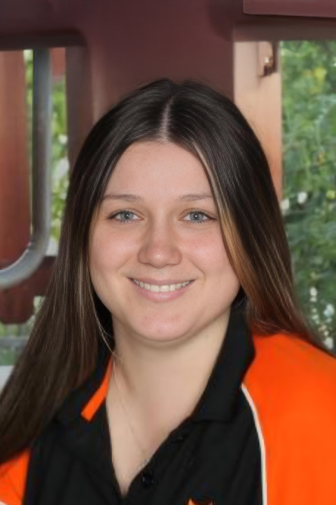 A headshot of Natteisha Gibson, smiling at the camera. She has long straight brown hair and is wearing a black and orange polo top.
