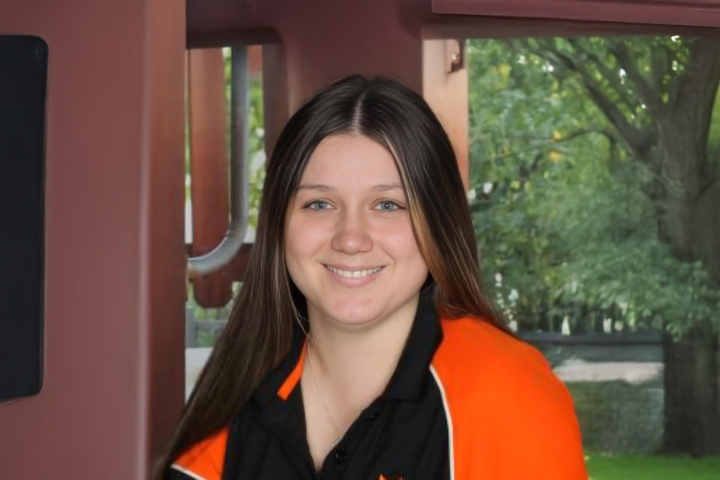 A headshot of Netteisha Gibson, smiling at the camera. She has long straight brown hair and is wearing a black and orange polo top.