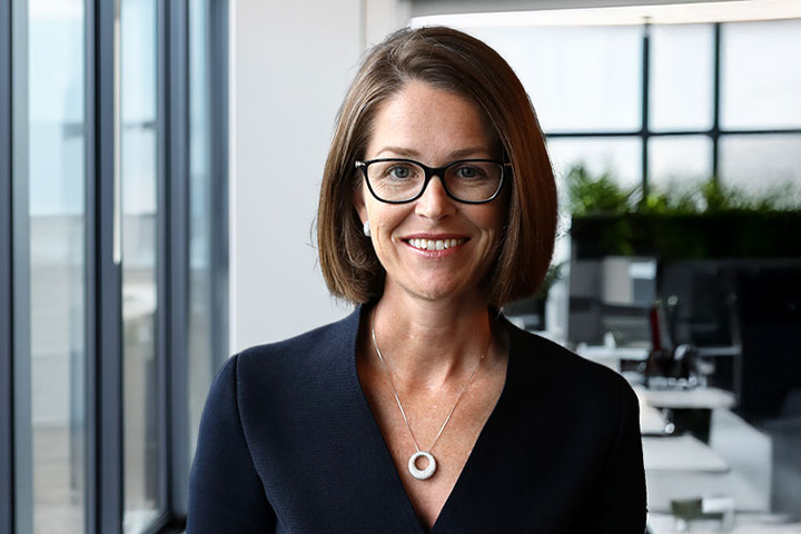 The image shows Alison Mirams in a professional and stylish black outfit. She stands with her hands behind her back and is smiling. She has shoulder length brown hair and is wearing glasses and a necklace.