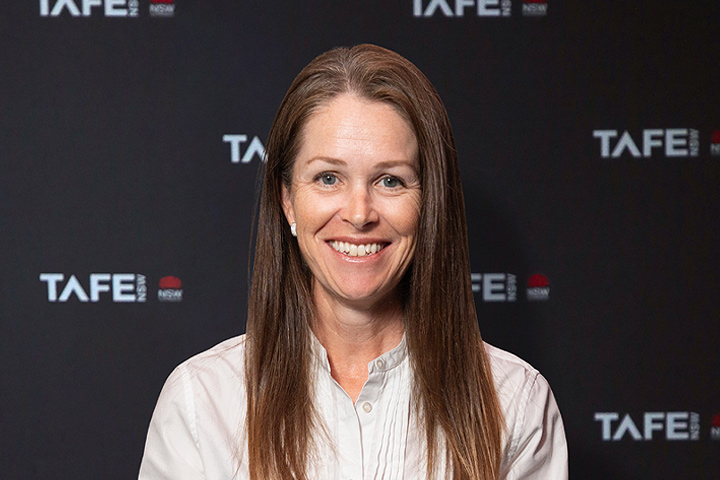 The image shows Alison Mirams in a professional and stylish black outfit. She stands with her hands behind her back and is smiling. She has shoulder length brown hair and is wearing glasses and a necklace.