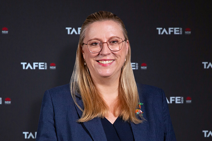 The image shows Chloe Read in a dark blue blazer standing in front of a TAFE NSW black backdrop. She has long blonde hair and is wearing glasses.
