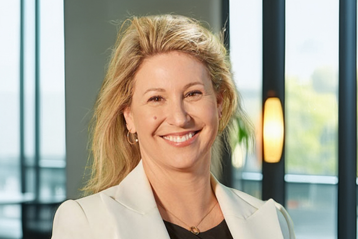 The image shows Eva Hanly smiling towards the camera wearing a white blazer won over a black top. She is wearing fine gold jewellery and has long blonde hair behind her shoulders.