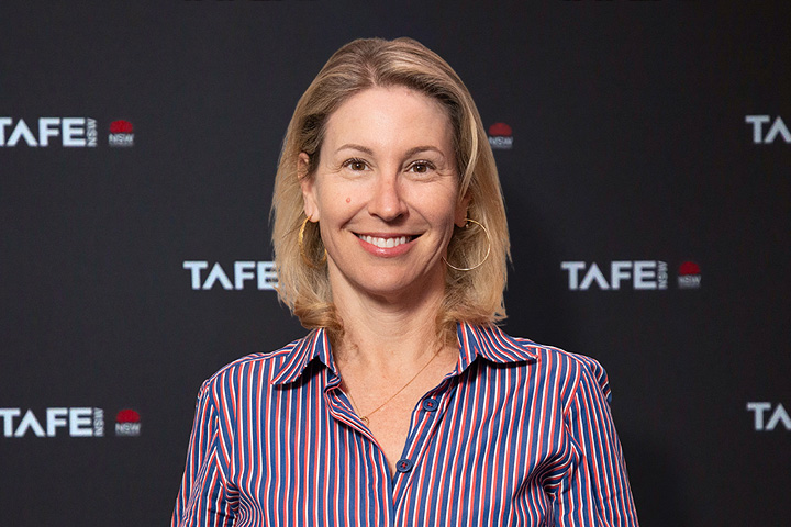 The image shows Eva Hanly smiling towards the camera wearing a white blazer won over a black top. She is wearing fine gold jewellery and has long blonde hair behind her shoulders.