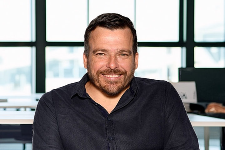 The image shows Kurt Fearnley in a black collared shirt. He has short brown hair and is smiling.