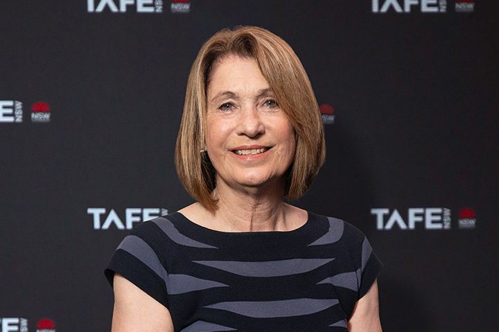 The image shows Mary Ann O’Loughlin up close. She's wearing a black jacket with a red shirt underneath. She has a short, light brown bob haircut and is smiling.