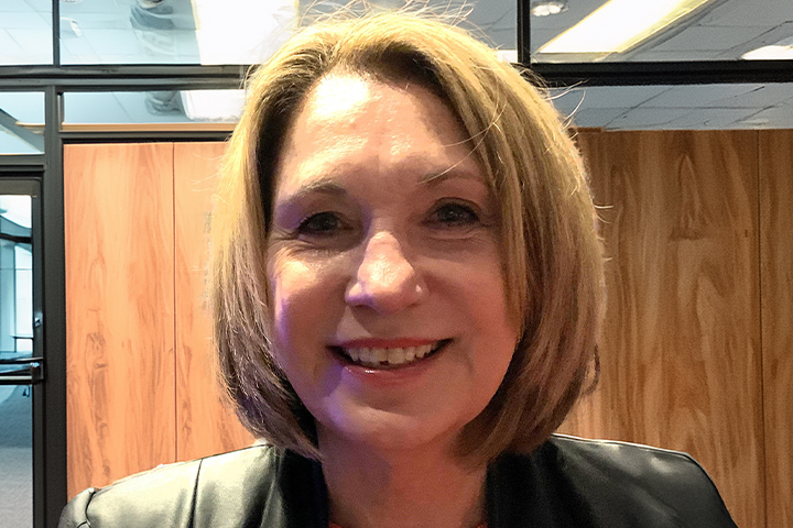 The image shows Mary Ann O’Loughlin up close. She's wearing a black jacket with a red shirt underneath. She has a short, light brown bob haircut and is smiling.