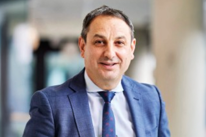 The image shows Murat Dizdar in a dark blue business suit, wearing a patterned blue tie. He has short hair and is smiling with his hands in his pockets.